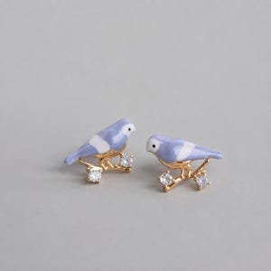 May include: A pair of stud earrings featuring gold-tone birds with light blue and white enamel detailing. Each bird is perched on a gold-tone branch accented with two small, clear, round-cut sparkling stones that resemble dew drops.