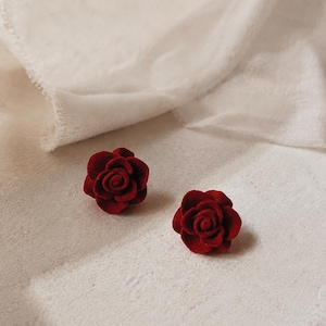 Op de afbeelding: Een paar oorknopjes in de vorm van dieprode rozen. Elke oorbel heeft een gelaagd, cirkelvormig bloembladontwerp met een matte, getextureerde afwerking die het uiterlijk van een bloeiende bloem nabootst. De rozen zijn klein, rond en symmetrisch.