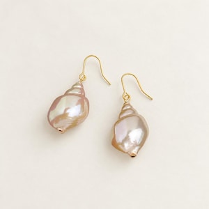 Peut inclure: Une paire de boucles d'oreilles pendantes composées de perles nacrées rose clair en forme de coquillage, suspendues à de fins crochets en métal doré. Les perles présentent une surface lisse et lustrée avec des reflets irisés.