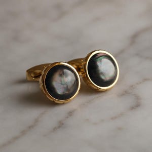 Può includere: Una coppia di gemelli rotondi con una montatura in metallo dorato lucido e un intarsio in madreperla scura iridescente. Le facce circolari mostrano un riflesso vorticoso e multicolore su uno sfondo nero profondo, con un bordo dentellato.
