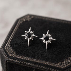 May include: A pair of silver-toned stud earrings featuring a central round black stone surrounded by a halo of small clear stones, with four elongated points extending outward in a starburst design, resting on a black velvet jewelry box.
