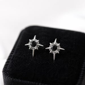 Puede incluir: Un par de pendientes de bot&oacute;n plateados con una piedra negra redonda central rodeada por un halo de peque&ntilde;as piedras claras, con cuatro puntas met&aacute;licas alargadas que crean una forma de estrella sobre una superficie de terciopelo negro.