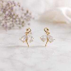 Könnte beinhalten: Ein Paar goldfarbene Ohrstecker in Form einer tanzenden Figur. Jeder Ohrring zeigt eine kleine goldene Metallsilhouette einer Person mit erhobenen Armen, akzentuiert durch zwei klare, tropfenförmige facettierte Steine als Rock.