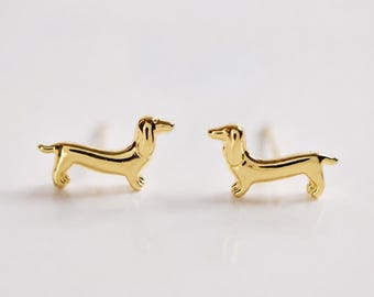 Op de afbeelding: Een paar goudkleurige oorknopjes in de vorm van teckels. Elke oorbel heeft een gladde, gepolijste metalen afwerking met het kenmerkende silhouet van de hond, inclusief een lang lichaam, korte pootjes en hangende oren, ontworpen als oorknopjes.