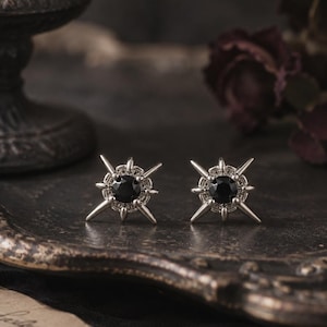 May include: A pair of silver-toned stud earrings featuring a central round black stone surrounded by a starburst design with multiple pointed rays extending outward from the center, creating a geometric and symmetrical celestial appearance.