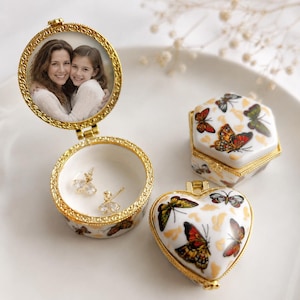 May include: Three small trinket boxes with gold-toned metal rims. One round box is open, displaying a photograph of two people inside the lid and two small stud earrings in the base. Two closed boxes are shaped like a hexagon and a heart.