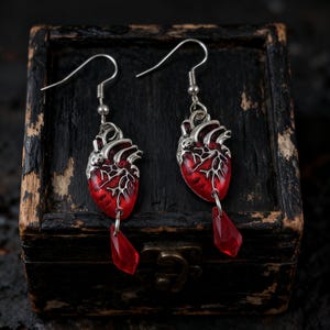 Könnte beinhalten: Silberfarbene Herzohrringe mit roter Emaille und einem hängenden roten Kristall. Die Ohrringe werden auf einer dunklen Holzkiste präsentiert. Gothic- oder alternativer Stil, ca. 3 cm lang.
