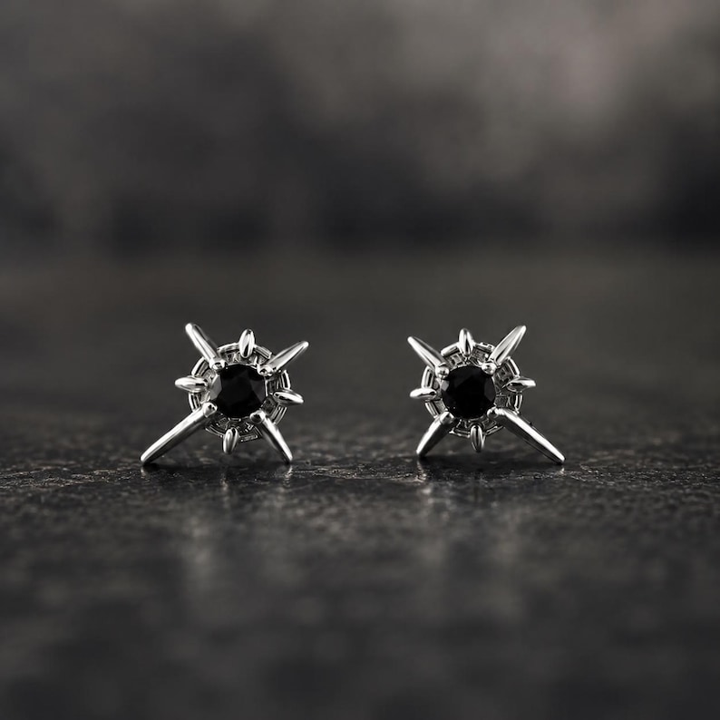 Puede incluir: Un par de pendientes de bot&oacute;n en tono plateado con una piedra negra redonda central rodeada por un dise&ntilde;o de estrella con m&uacute;ltiples puntas met&aacute;licas radiantes. Los pendientes tienen un acabado met&aacute;lico y una forma sim&eacute;trica.