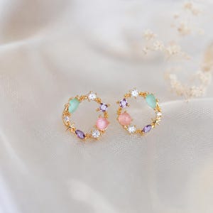 Op de afbeelding: Een paar goudkleurige, ronde oorknopjes met een delicaat kransontwerp. Elke oorbel is versierd met kleine heldere kristallen, ronde roze en mintgroene cabochons, paarse stenen in marquise-slijpvorm en kleine gouden stervormige accenten.