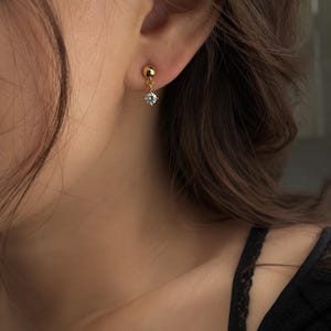 May include: A gold-coloured earring featuring a small, round stud and a dangling, clear, round gemstone. The earring is worn on the ear of a person with brown hair. The person is wearing a black top.