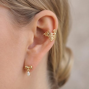 Pode incluir: Um ear cuff dourado com um design floral delicado e vazado, acompanhado por um brinco de pino dourado em forma de laço pequeno com uma pedra transparente em formato de gota pendurada, usados no lóbulo e na cartilagem da orelha.