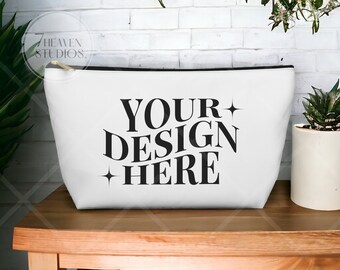 May include: A white cosmetic bag with a black zipper and the text "YOUR DESIGN HERE" in bold black letters. The bag is sitting on a wooden table. A small succulent plant in a white pot is to the right.