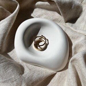 Può includere: Un porta anelli in ceramica bianca con una forma organica unica. Due anelli d'oro sono appoggiati sul piatto.