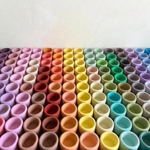 Könnte beinhalten: Eine Anordnung kleiner, zylindrischer Objekte in einem Regenbogenfarbverlauf. Die Objekte sind in verschiedenen Farbtönen bemalt, von Rosa bis Blau, wodurch ein visuell ansprechendes Muster entsteht. Die Objekte sind gleichmäßig angeordnet und bilden ein farbenfrohes Arrangement.