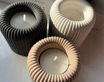 May include: Three concrete candle holders in black, white, and beige with a ribbed design. Each holder has a small tea light candle inside.