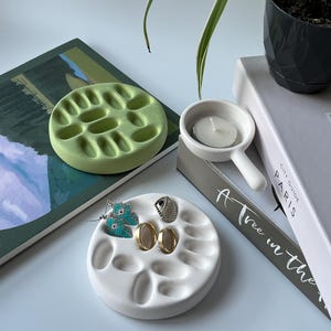 May include: A white ceramic dish with multiple indentations for holding jewelry. The dish is filled with earrings and rings. The dish is sitting on a book with the text "A Tree in the City Guide Paris".
