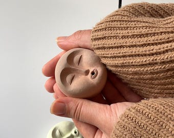 May include: A terracotta-colored ceramic incense holder shaped like a face with closed eyes and a small hole for incense. The holder is being held in a hand wearing a brown sweater.