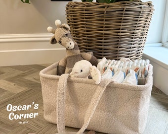 May include: A beige fuzzy storage basket with two handles, filled with diapers, a stuffed giraffe, and a stuffed bunny. The basket is sitting on a wood floor with a wicker basket and a plant in the background. The text "Oscar's Corner Est. 2011" is visible below the basket.