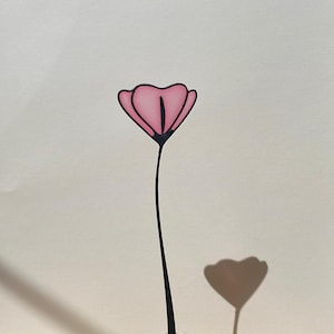 Puede incluir: Una escultura de flor rosa con tallo y base negros. La flor tiene un diseño en forma de corazón. La escultura está sobre una superficie de madera con fondo blanco. La sombra de la flor es visible.