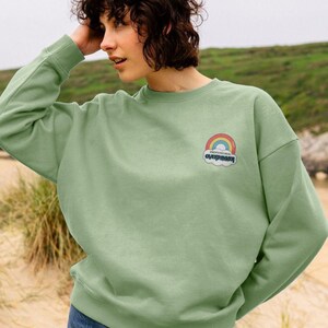 Puede incluir: Una sudadera de color verde claro con un gráfico de arcoíris y nube que dice "Professional Overthinker".