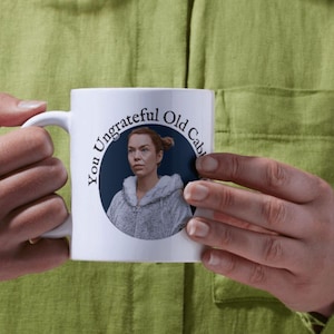 May include: White ceramic mug with a blue circle and the text "You Ungrateful Old Cable" in black. A woman with brown hair is pictured inside the circle.