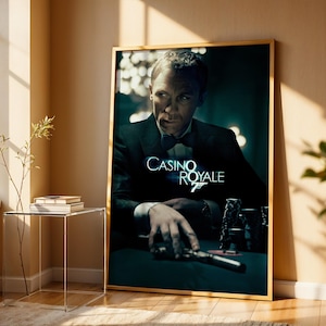 May include: A framed movie poster for Casino Royale. The poster features a man in a tuxedo, with the film's title in white lettering. The poster is in a gold frame and is leaning against a wall.