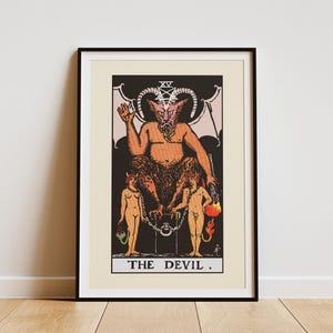 Peut inclure: Impression encadrée de carte de tarot représentant le Diable. L'image montre une figure cornue à la peau orange assise sur un trône, avec deux figures nues enchaînées en dessous. La carte est intitulée "THE DEVIL" en bas.