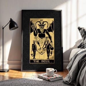Peut inclure: Impression d'art encadrée noire avec une illustration dorée et noire de la carte de tarot du Diable. L'œuvre représente une figure cornue avec des ailes de chauve-souris, entourée de deux figures nues. Le texte "THE DEVIL" est en bas.