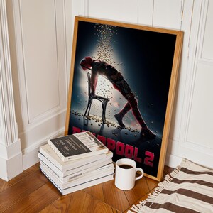 May include: A framed poster for the movie Deadpool 2, featuring a figure in a red suit leaning on a chair. The poster is in a wooden frame and is propped against a white wall. A stack of books and a coffee mug are in front of the poster.