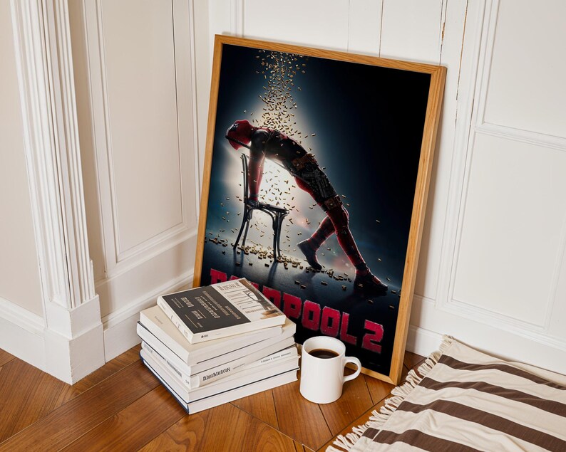 May include: A framed poster for the movie Deadpool 2, featuring a figure in a red suit leaning on a chair. The poster is in a wooden frame and is propped against a white wall. A stack of books and a coffee mug are in front of the poster.