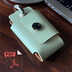 May include: A light green leather key holder with a silver-tone clasp and a gold-colored snap closure. The key holder is rectangular with a folded flap. The background is a dark wood surface.
