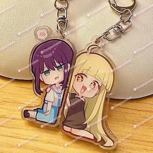Puede incluir: Dos llaveros de acrílico con personajes de anime estilo chibi. Un personaje tiene cabello morado oscuro y viste una blusa marinera blanca, mientras que el otro tiene cabello largo rubio y viste un vestido marrón oscuro. Incluyen anillas de metal.