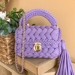 May include: A lavender crochet handbag featuring a thick braided texture, a sturdy top handle, and a gold-toned metal push-lock closure. The bag includes a matching lavender crochet and gold chain shoulder strap and a large decorative tassel.