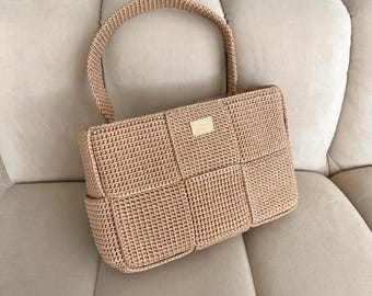 Puede incluir: Un bolso de mano de ganchillo beige con un patrón de cuadrados tejidos y una sola asa superior. El bolso tiene una estructura rectangular con una pequeña placa metálica rectangular de tono dorado centrada en el panel frontal, con texto grabado.