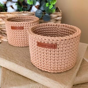 May include: Two round, light brown crochet baskets made from thick cord. Each basket features a small, rectangular brown leather-like tag on the front with the word "Handmade" embossed in a dark brown font, centered on the textured knit surface.