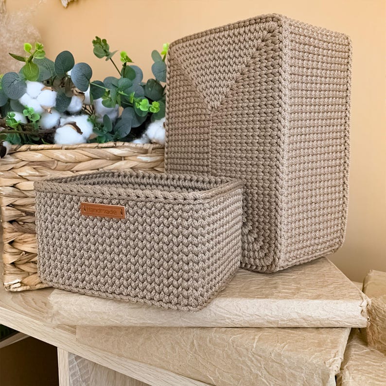 Op de afbeelding: Twee rechthoekige opbergmanden gemaakt van beige gehaakt koord. De kleinere mand aan de voorzijde is voorzien van een rechthoekig bruin leren label met het woord "handmade" erin gedrukt. De manden hebben een consistente, dichte, textuursteek.