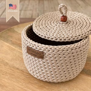 May include: A beige crocheted storage basket with a lid and a wooden knob. The basket has a brown leather label that says "WOWBAG EXCLUSIVE".