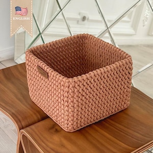 May include: A square-shaped, woven storage basket made of a light brown, textured material. The basket is empty and sitting on a wooden surface.