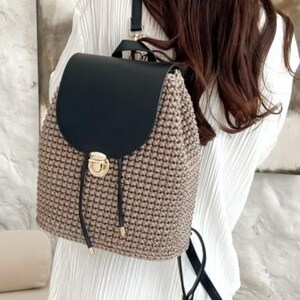 May include: A beige and black backpack with a flap closure. The backpack features a woven design on the main body and a black flap. It has a drawstring closure with a gold-colored clasp and black straps. The bag is designed to be worn on the back.