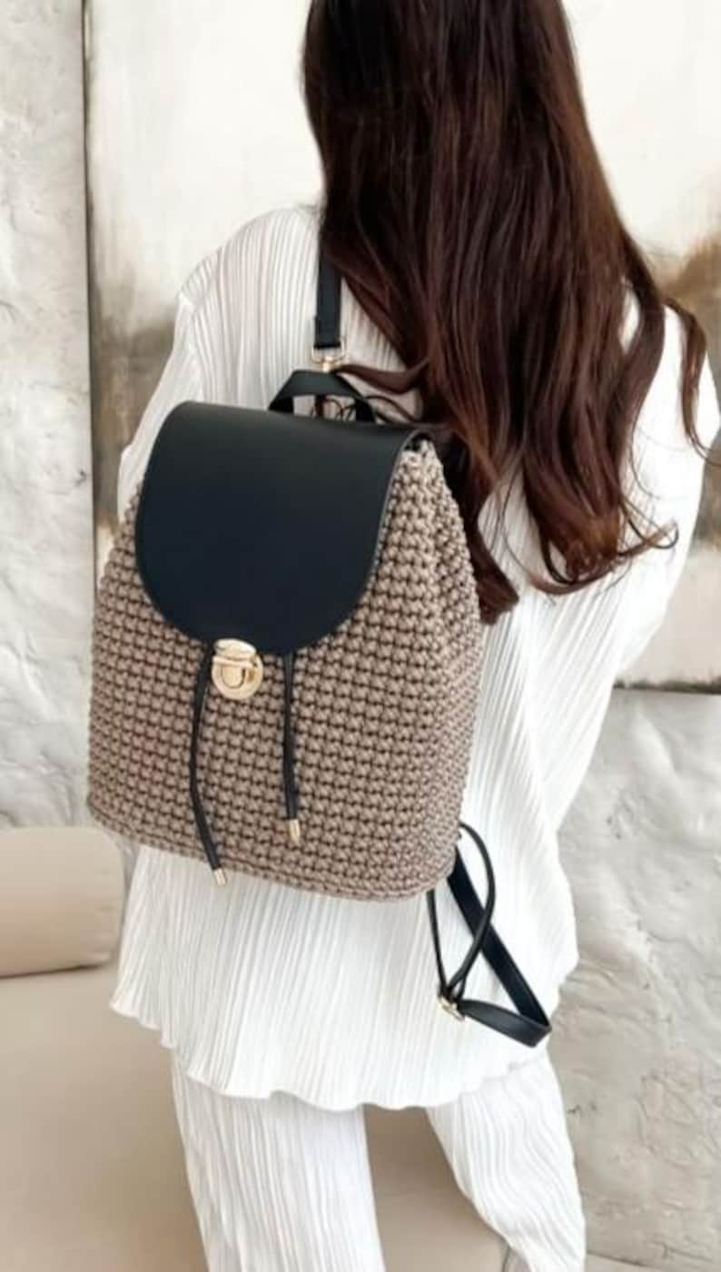 May include: A beige and black backpack with a flap closure. The backpack features a woven design on the main body and a black flap. It has a drawstring closure with a gold-colored clasp and black straps. The bag is designed to be worn on the back.