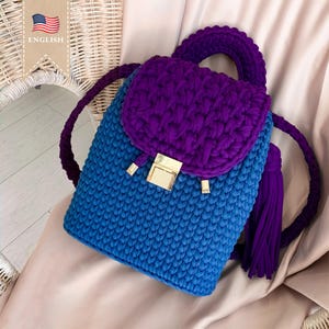 May include: A small, crocheted backpack with a purple top and a blue bottom. The backpack has a gold clasp and a purple tassel.
