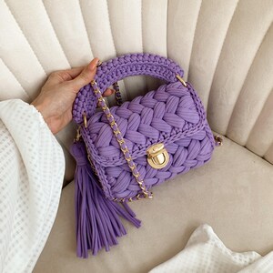 May include: A lavender purple handbag crafted from thick braided fabric, featuring a gold-toned metal push-lock closure, a matching braided top handle, a long gold chain shoulder strap, and a large decorative tassel hanging from the side.