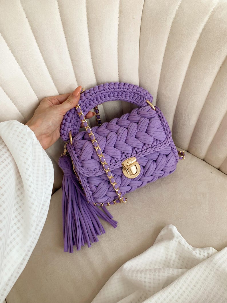 May include: A lavender purple handbag crafted from thick braided fabric, featuring a gold-toned metal push-lock closure, a matching braided top handle, a long gold chain shoulder strap, and a large decorative tassel hanging from the side.