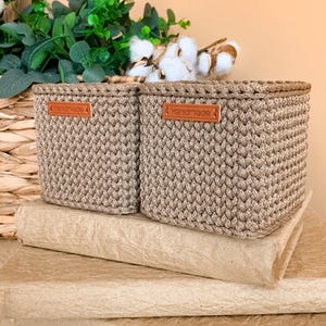 Könnte beinhalten: Zwei quadratische, beige gehäkelte Aufbewahrungskörbe mit strukturiertem Strickmuster. Jeder Korb hat vorne ein rechteckiges, braunes lederartiges Etikett, auf dem das Wort "Handmade" in heller Farbe im oberen Bereich eingeprägt ist.