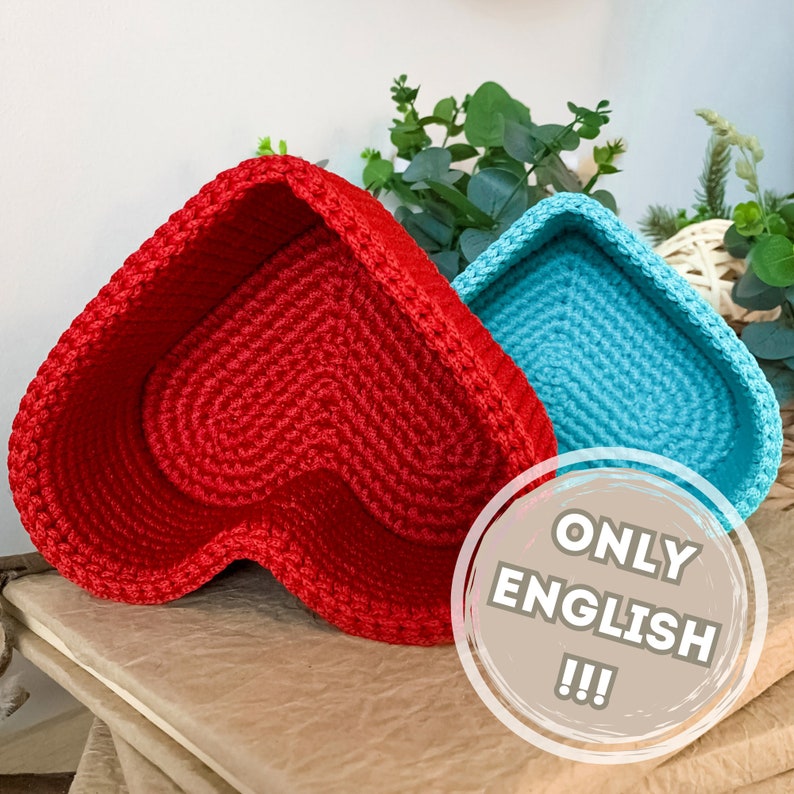 May include: Two crocheted heart-shaped baskets, one red and one blue. The red basket is in the foreground and the blue basket is behind it. The baskets are sitting on a wooden surface.