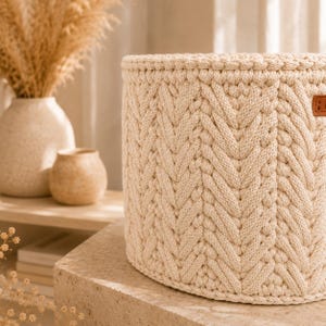 Puede incluir: Una cesta de almacenamiento cil&iacute;ndrica hecha de cuerda gruesa de color crema con un patr&oacute;n de tejido de espiga. La cesta incluye una peque&ntilde;a etiqueta rectangular de color marr&oacute;n con aspecto de cuero en el borde superior lateral.