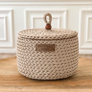 May include: A round, beige, crocheted storage basket with a matching lid. The lid features a central loop handle topped with a small, dark brown wooden bead. A rectangular brown leather-like label on the front displays the text WOWBAG EXCLUSIVE.