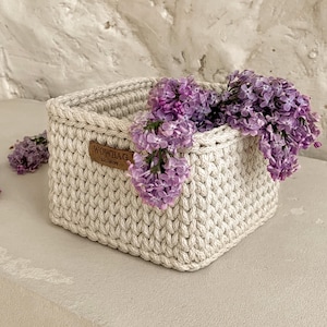 May include: A square, off-white crocheted basket with a thick, textured cord stitch. A brown leather-like tag with the text WOWBAG EXCLUSIVE is attached to the side. The basket is filled with clusters of vibrant purple lilac flowers.