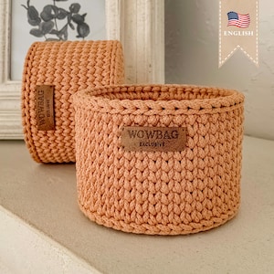 May include: Two peach-colored, hand-crocheted baskets. The larger basket is round, while the smaller one is rectangular. Both have a woven texture and a brown leather tag that reads "WOWBAG EXCLUSIVE". The baskets are displayed on a white surface.