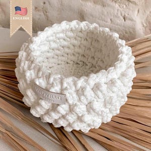 May include: White crocheted basket with a woven texture. The basket is sitting on a light brown surface. The label on the basket reads "WOWBAG Creative."